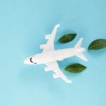 White airplane model emitting fresh green leaves on blue background. Sustainable travel; clean and green energy; and biofuel for aviation industry concept.