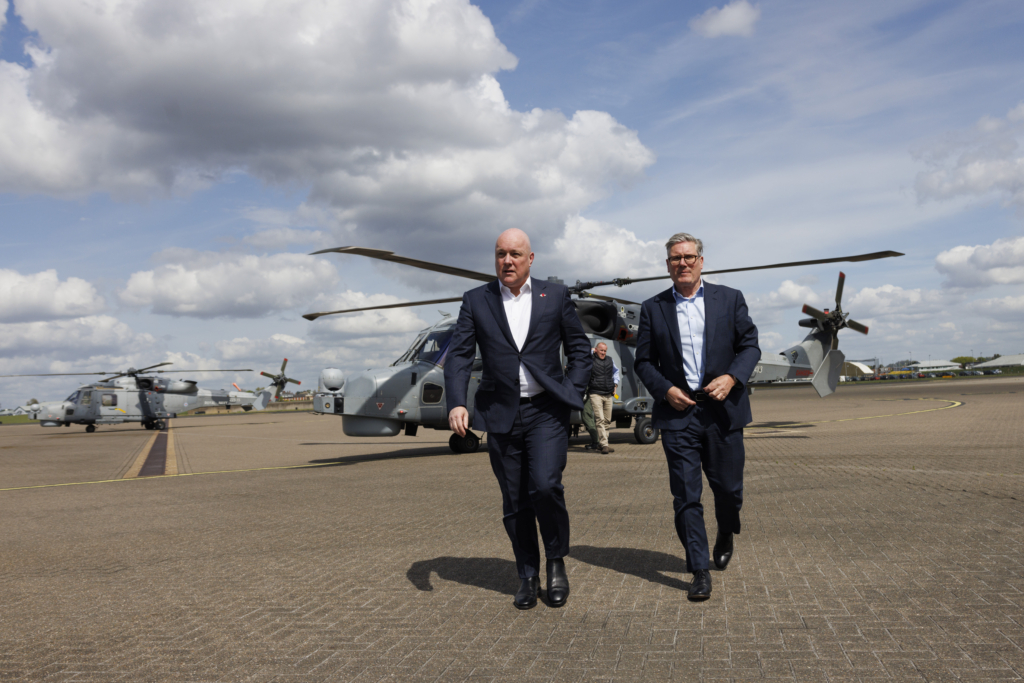 Two men in business suits walk across an airport tarmac with military helicopters in the background under a cloudy sky. The scene appears to be from an official government or military visit.