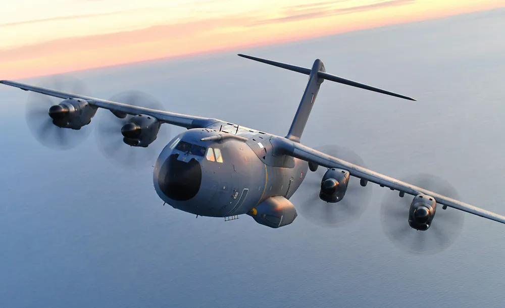 A large military transport aircraft flies through cloudy skies at sunset, with its four turboprop engines visible and the cockpit windows illuminated against the dramatic pink and orange evening sky.