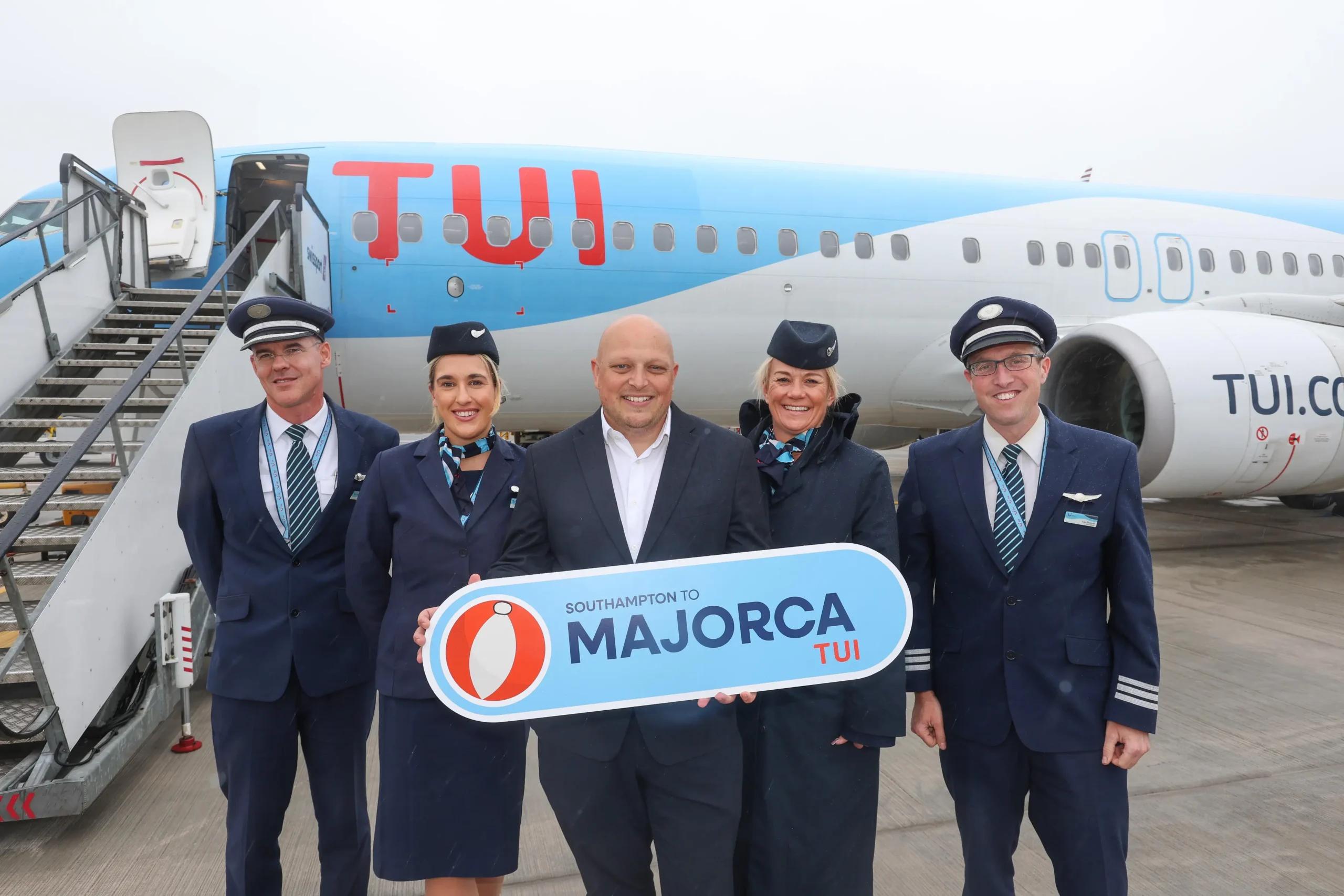 Five airline crew members in blue uniforms pose in front of a TUI aircraft, holding a sign that reads 