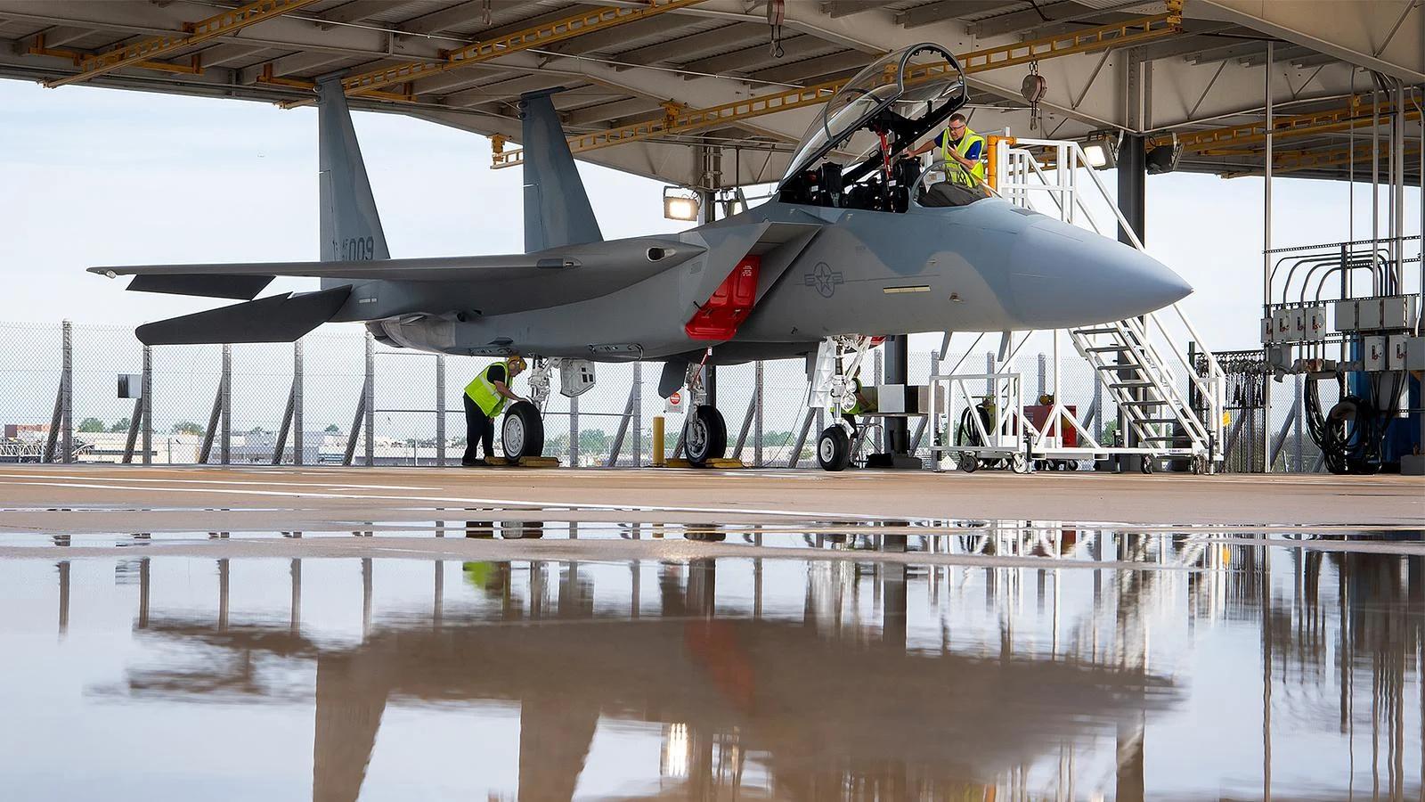 A military fighter jet undergoes maintenance in an aircraft hangar, with ground crew workers in high-visibility vests servicing the aircraft while it's positioned on a reflective wet floor that mirrors the scene above.