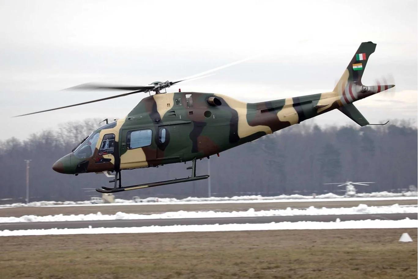 A military helicopter with green and tan camouflage paint hovers low over a snow-covered airfield during winter conditions. The aircraft appears to be performing a landing approach with bare trees visible in the background.