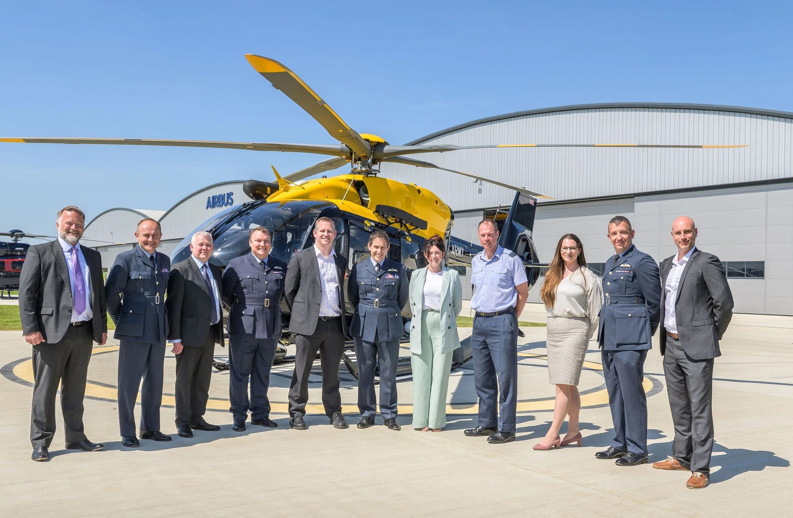 A group of approximately twelve people in business attire and military uniforms pose together in front of a yellow helicopter on an airport tarmac, with an aircraft hangar visible in the background.