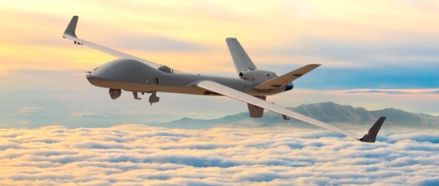 A military drone flies above a thick layer of clouds with mountains visible in the distance under a colorful sunset sky with orange and pink hues.