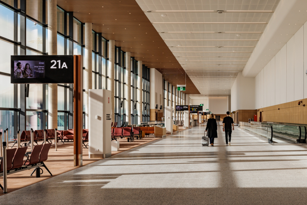 A modern airport terminal interior showing gate 21A with rows of red seating, large windows, and two travelers walking down a bright, spacious corridor with wood and white ceiling details.