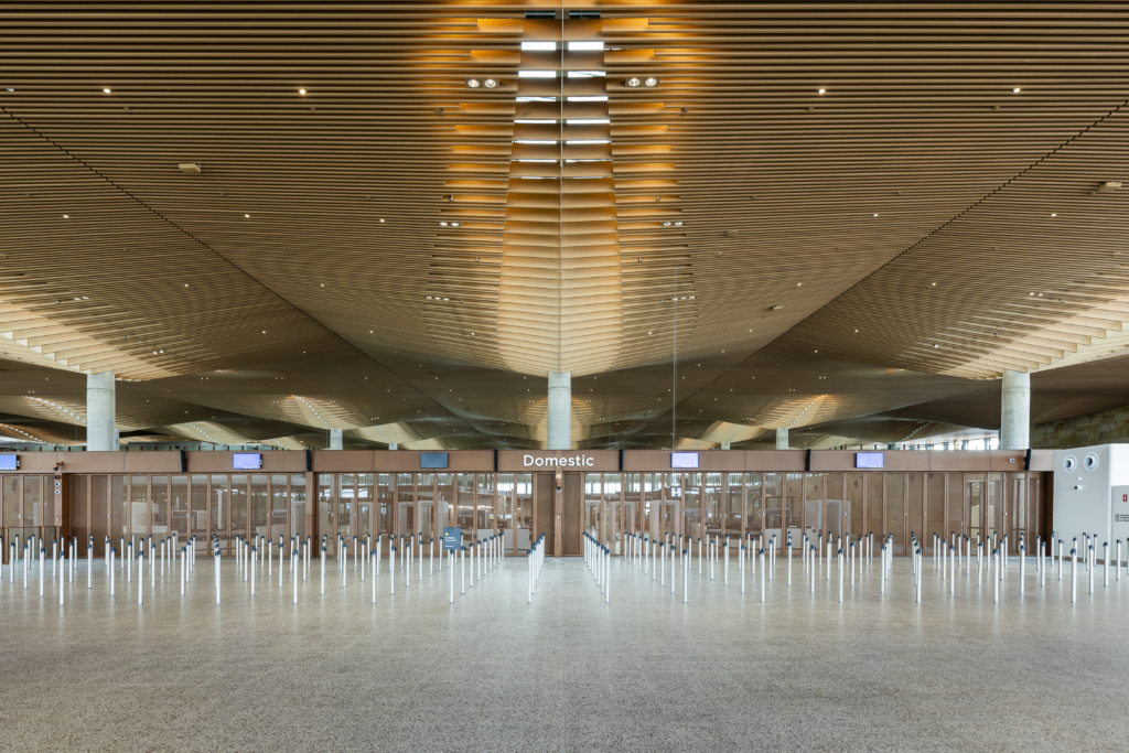 A modern airport terminal interior featuring a distinctive wooden slatted ceiling with dramatic lighting, rows of security barriers leading to domestic gates, white support columns, and polished concrete flooring creating a spacious, contemporary architectural design.