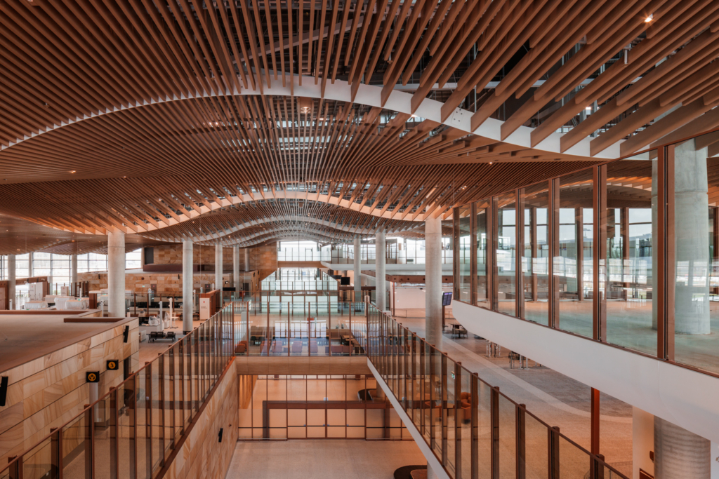 A modern interior space features a distinctive curved wooden slat ceiling that creates flowing, wave-like patterns above multiple levels connected by staircases and walkways. The warm wood tones complement the open, airy architecture with large windows and clean lines throughout the multi-story building.