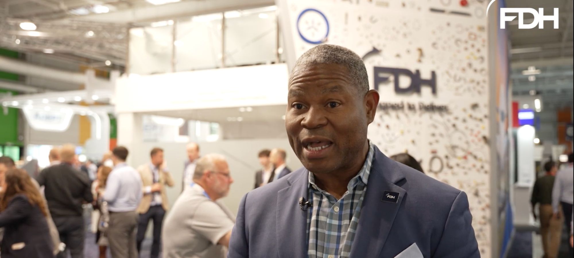 A professional man in a blue suit jacket and checkered shirt speaks at what appears to be a trade show or conference, with FDH signage and attendees visible in the background.