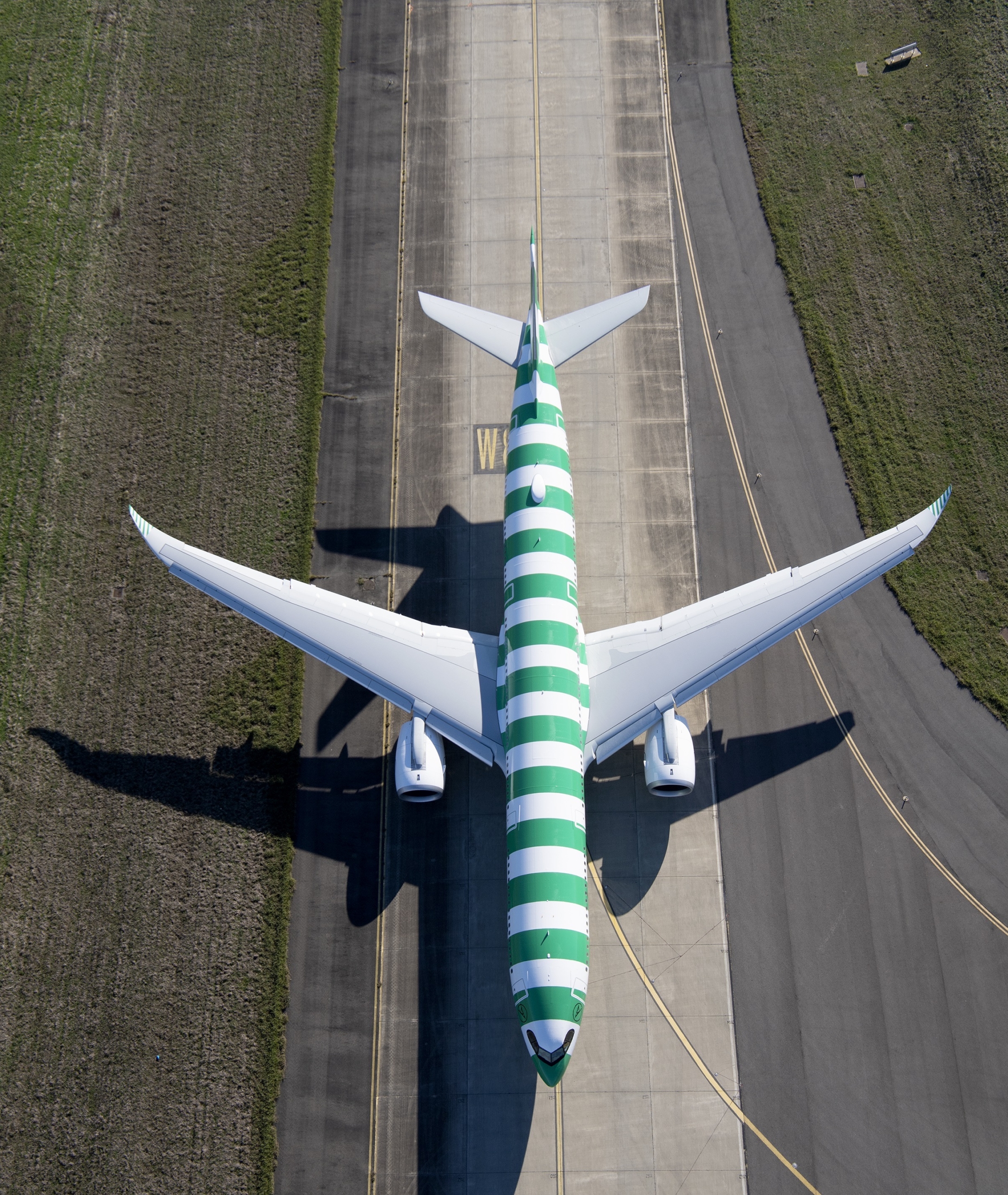 An aerial view of a commercial aircraft with distinctive green and white striped livery positioned on an airport runway, casting a clear shadow on the concrete surface with grass visible on either side.