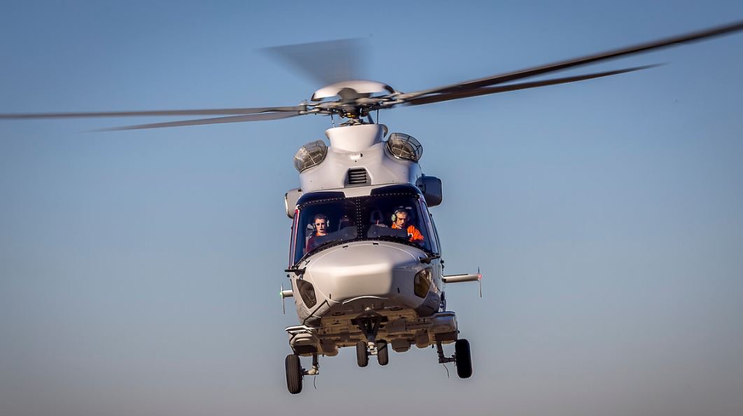 A white helicopter flies through clear blue skies with two pilots visible in the cockpit, its main rotor blades spinning overhead.