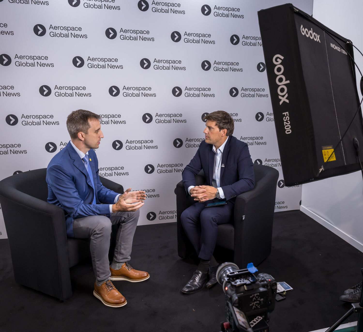 Two men in business suits sit facing each other on black chairs during what appears to be a filmed interview, with professional lighting equipment visible and an 
