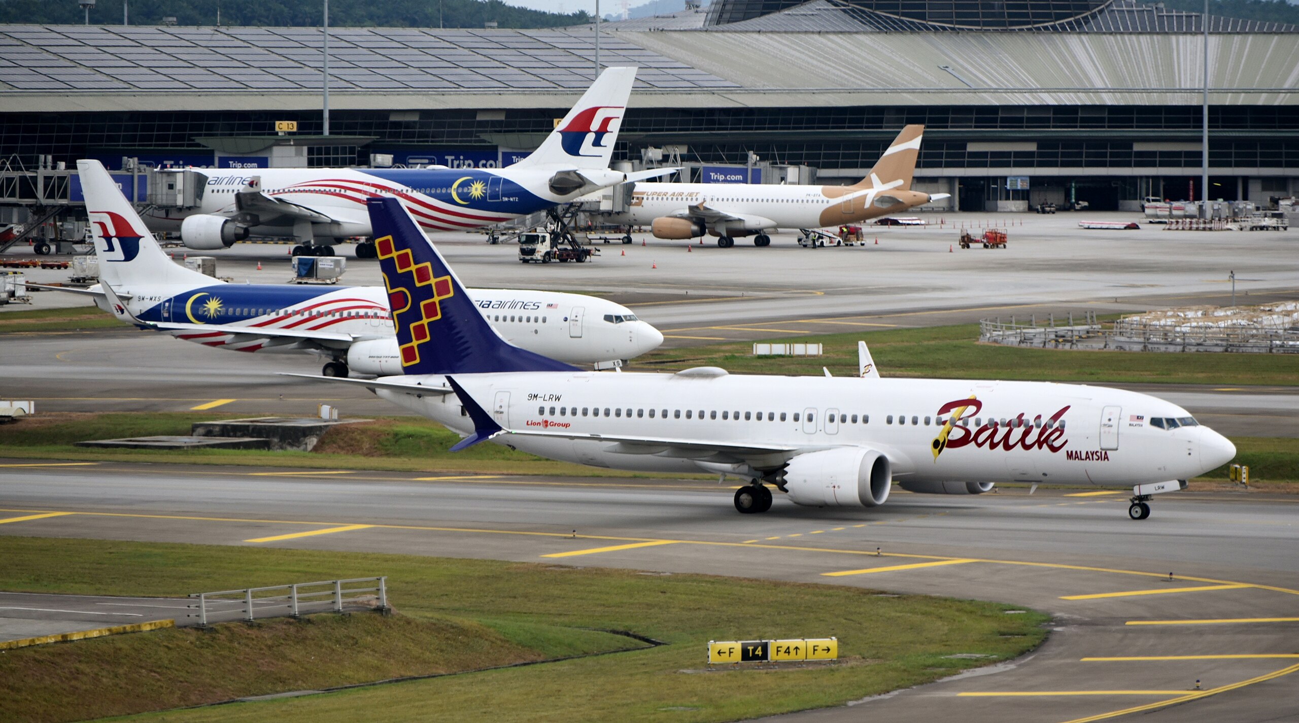 Several commercial aircraft are parked at gates and taxiing on runways at a busy airport, including planes from Malaysia Airlines, Batik Air Malaysia, and other carriers with distinctive liveries featuring Malaysian flag colors and designs.