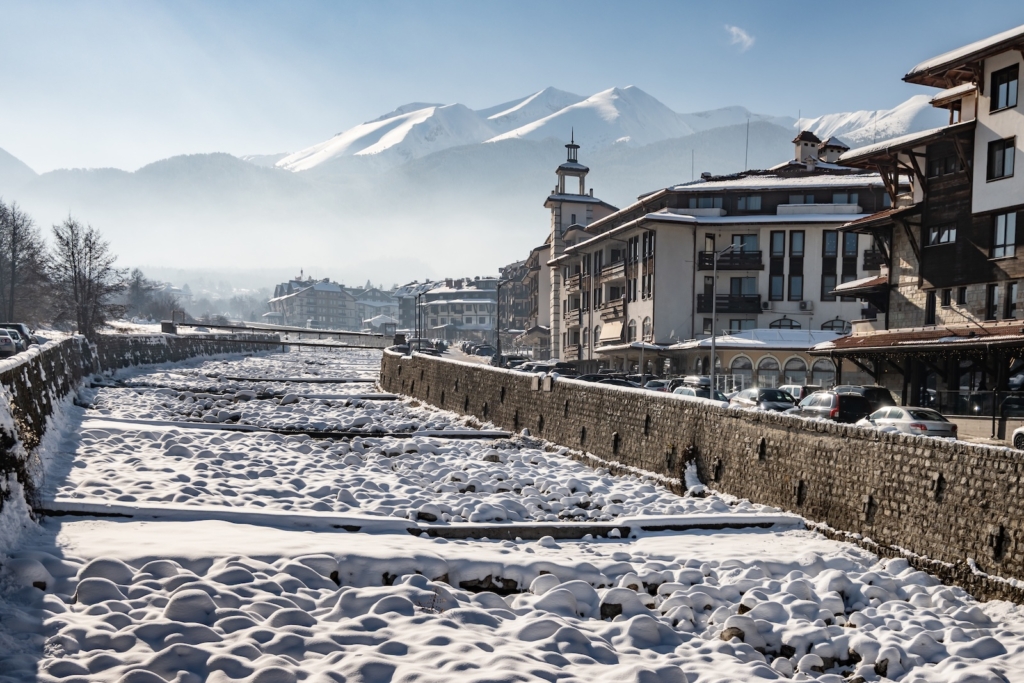 A frozen Glazne riverbed with snow-capped boulders creates a textured foreground against a stone wall, buildings, and the Pirin mountain range in Bansko, Bulgaria.