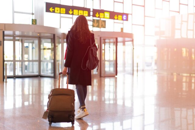 Rear view of brunette woman exit from airport with trolley (hand luggage)