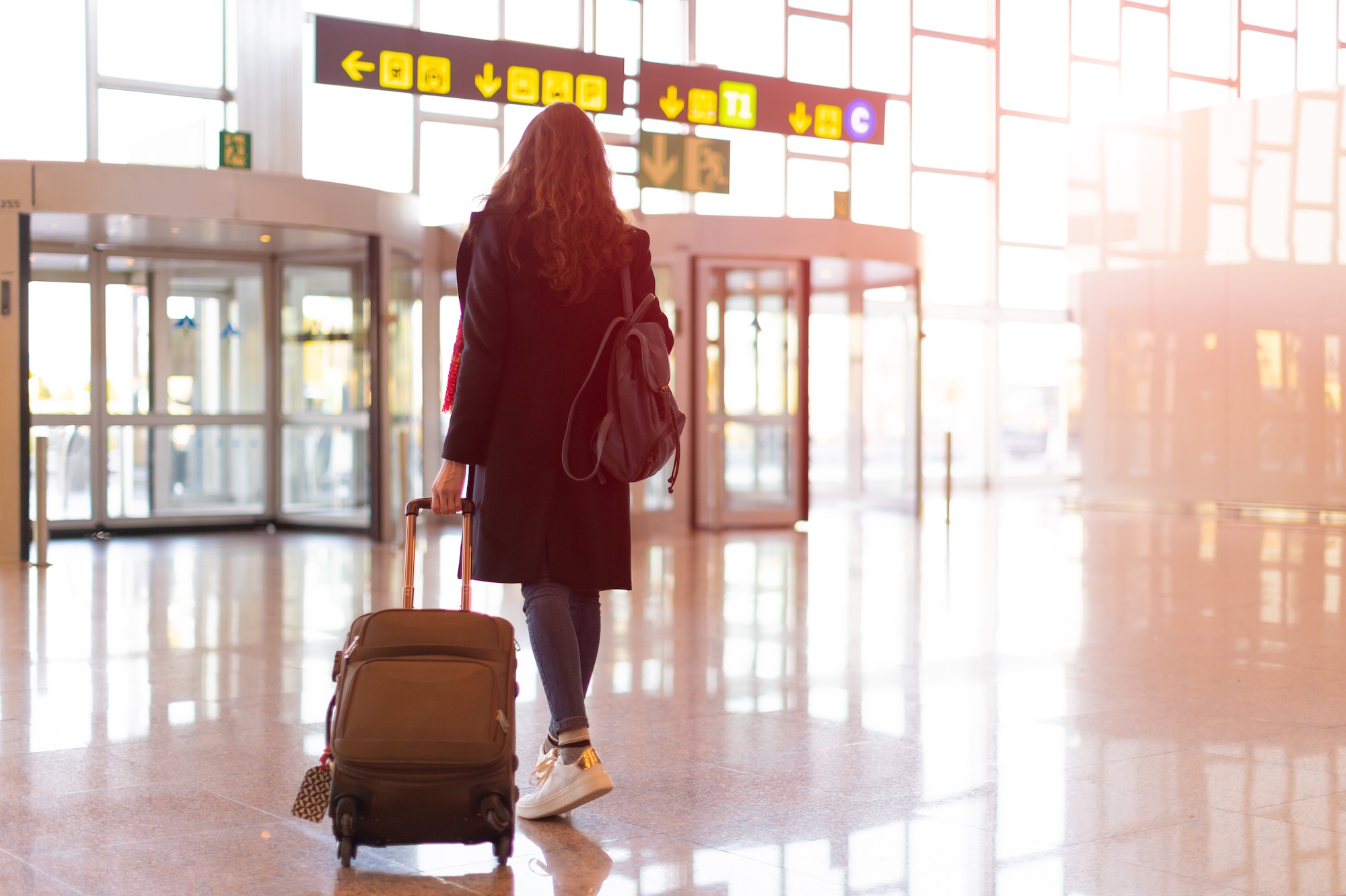 Rear view of brunette woman exit from airport with trolley (hand luggage)