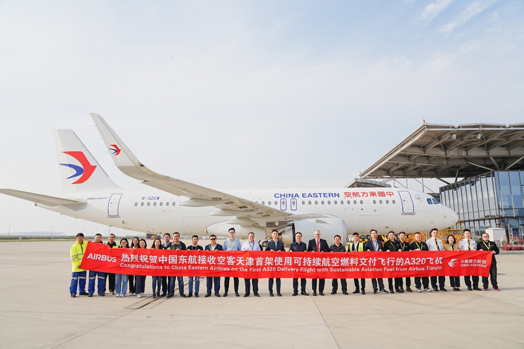 A group of officials and dignitaries hold a red ceremonial banner in front of a China Eastern Airlines Airbus A320 aircraft at an airport, celebrating the delivery of the airline's first A320 flight using sustainable aviation fuel.