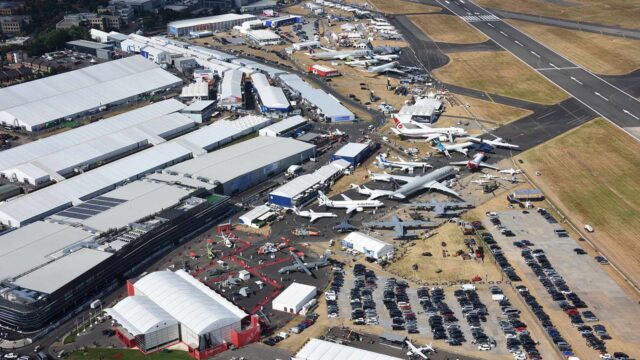 Aerial view of an airport showing multiple aircraft parked on the tarmac, large terminal buildings with white roofs, runways, and extensive parking lots filled with cars.