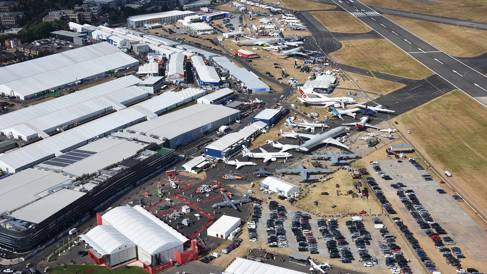 Aerial view of an airport showing multiple aircraft parked on the tarmac, large terminal buildings with white roofs, runways, and extensive parking lots filled with cars.