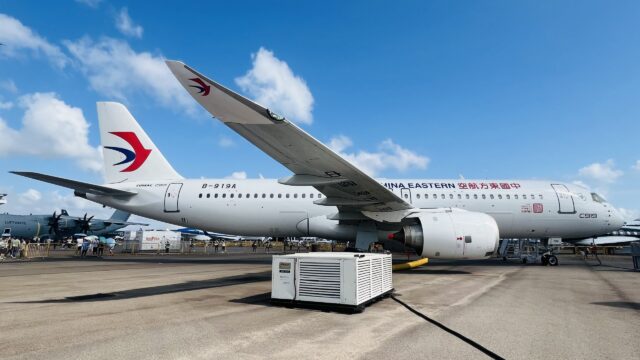 A China Eastern Airlines aircraft parked on an airport tarmac with ground support equipment nearby, displaying the airline's distinctive red and blue logo on its white fuselage under a partly cloudy blue sky.