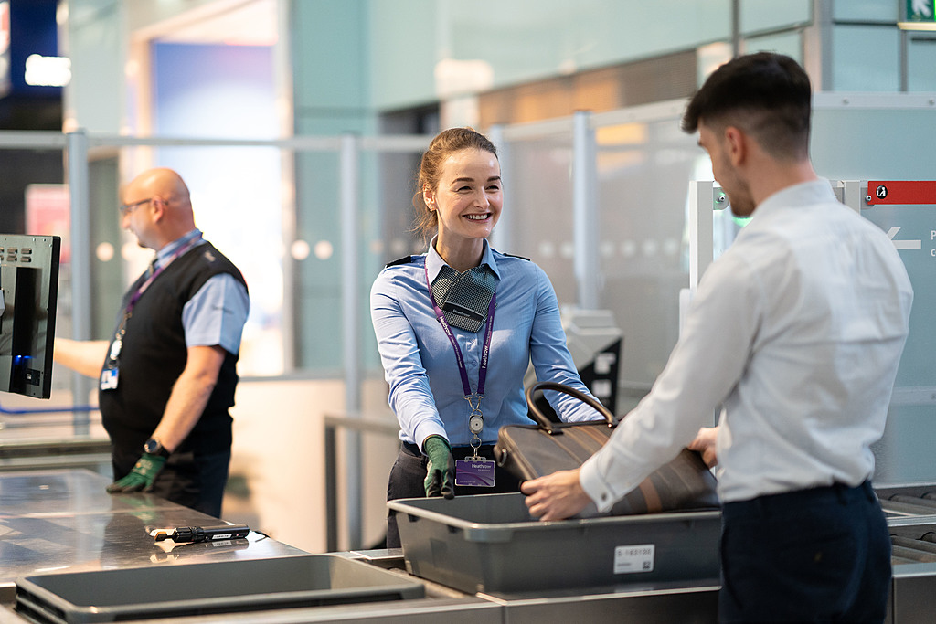 T5 Security performing different tasks. Security staff help passengers with checks on personal belongings in trays at a security area in Terminal 5 at London Heathrow Airport.