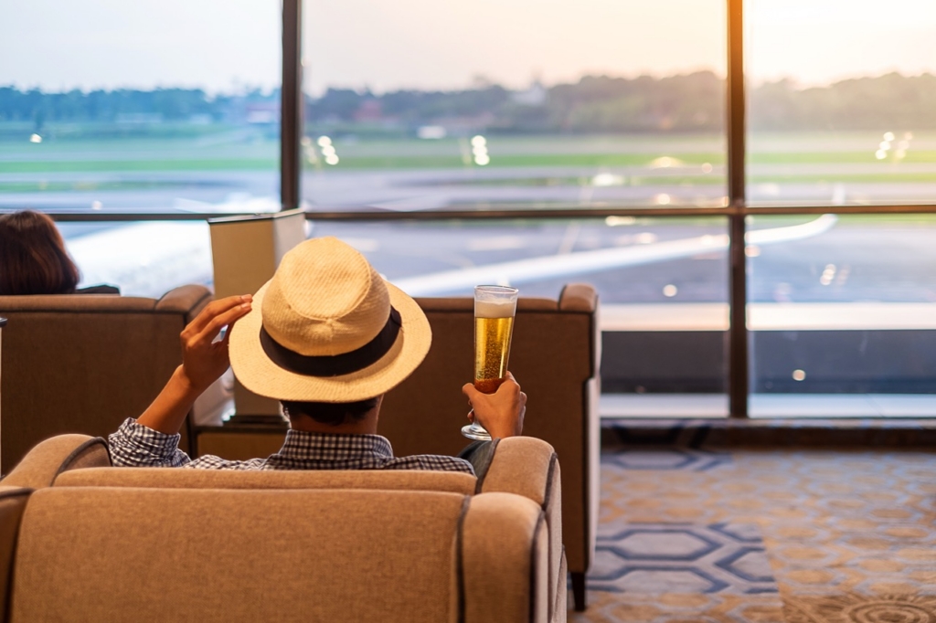 man traveler with hat holding beer glass and looking to airplane, Asian passenger sitting and relax in modern lounge at international airport terminal. 