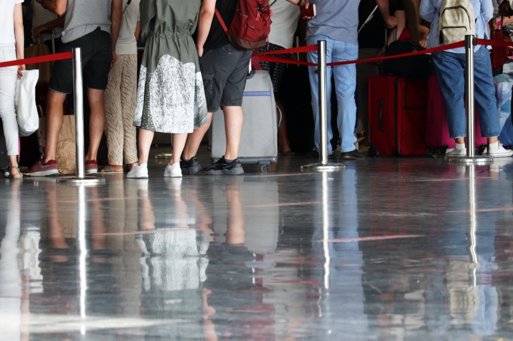 People with luggage standing in a queue behind red rope barriers on a highly polished reflective floor, likely at an airport or similar transit facility.