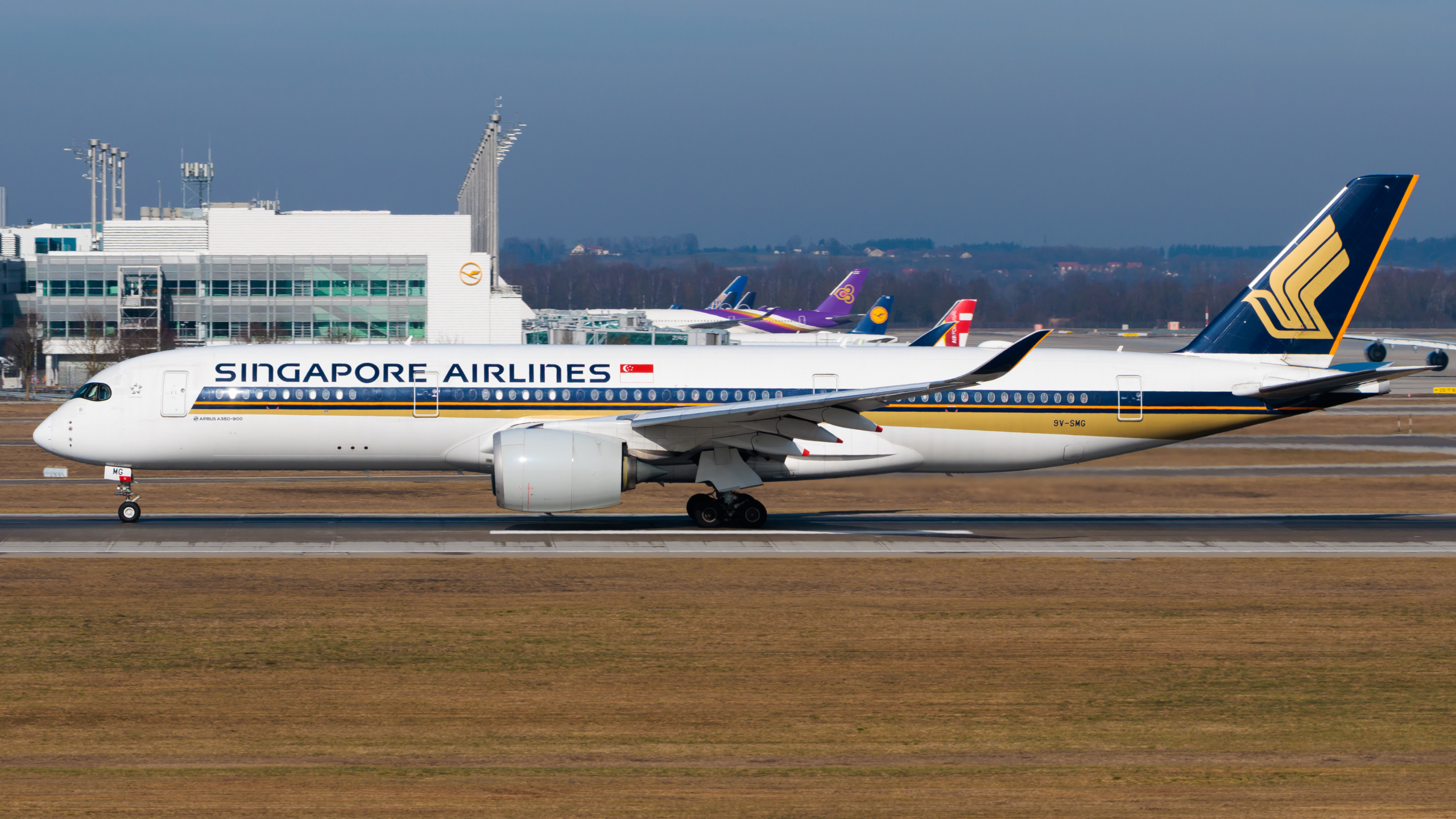 A Singapore Airlines aircraft with distinctive gold and blue livery sits on an airport runway, with the airline's logo visible on the white fuselage and terminal buildings in the background.