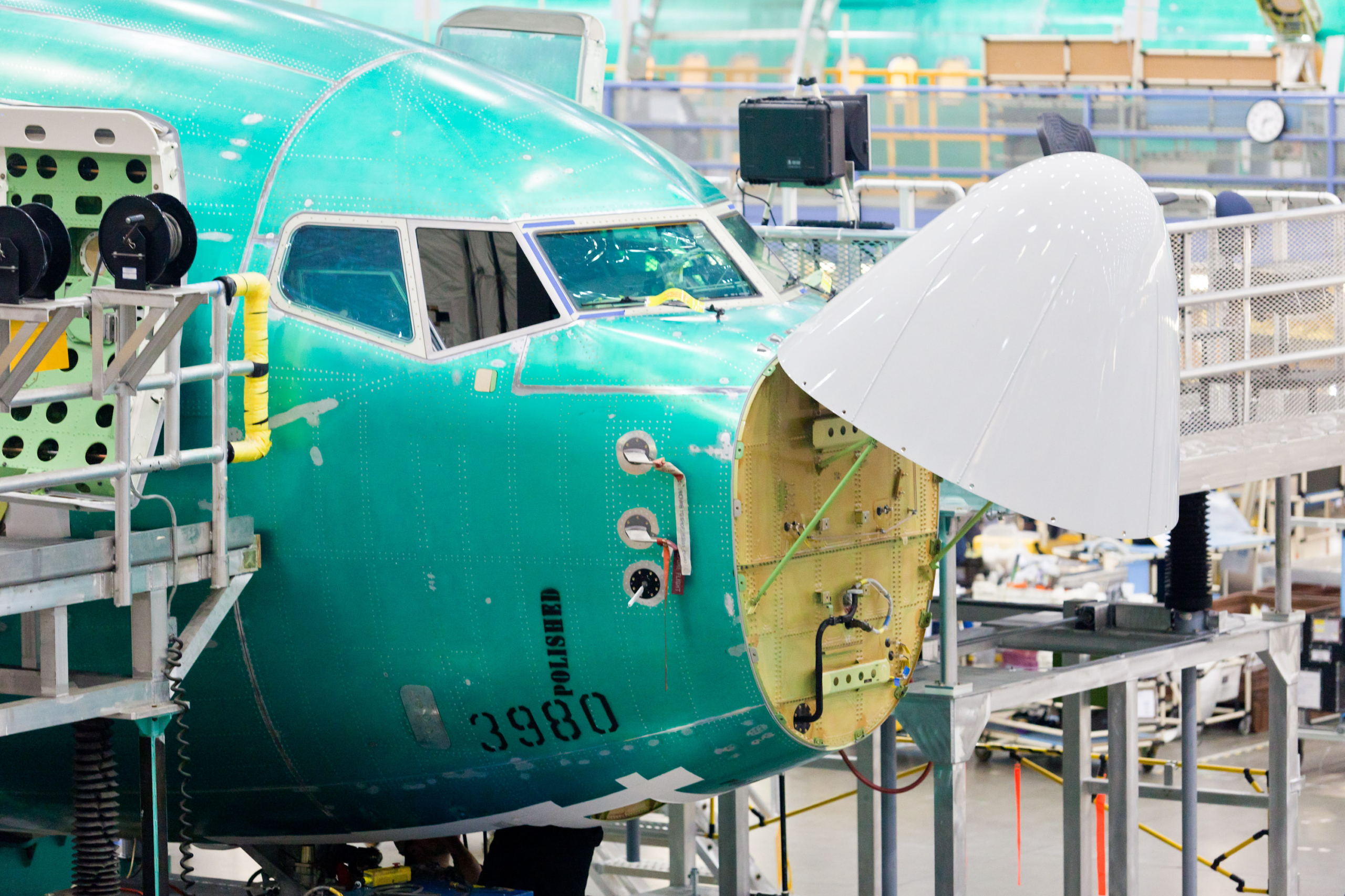 Aircraft fuselage section in green primer paint undergoing assembly in a manufacturing facility, with scaffolding and industrial equipment surrounding the nose area where internal components are visible.