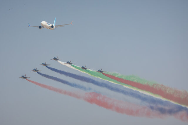 Aircraft performing aerial display at Dubai Airshow with colored smoke trails and commercial airplane flying overhead