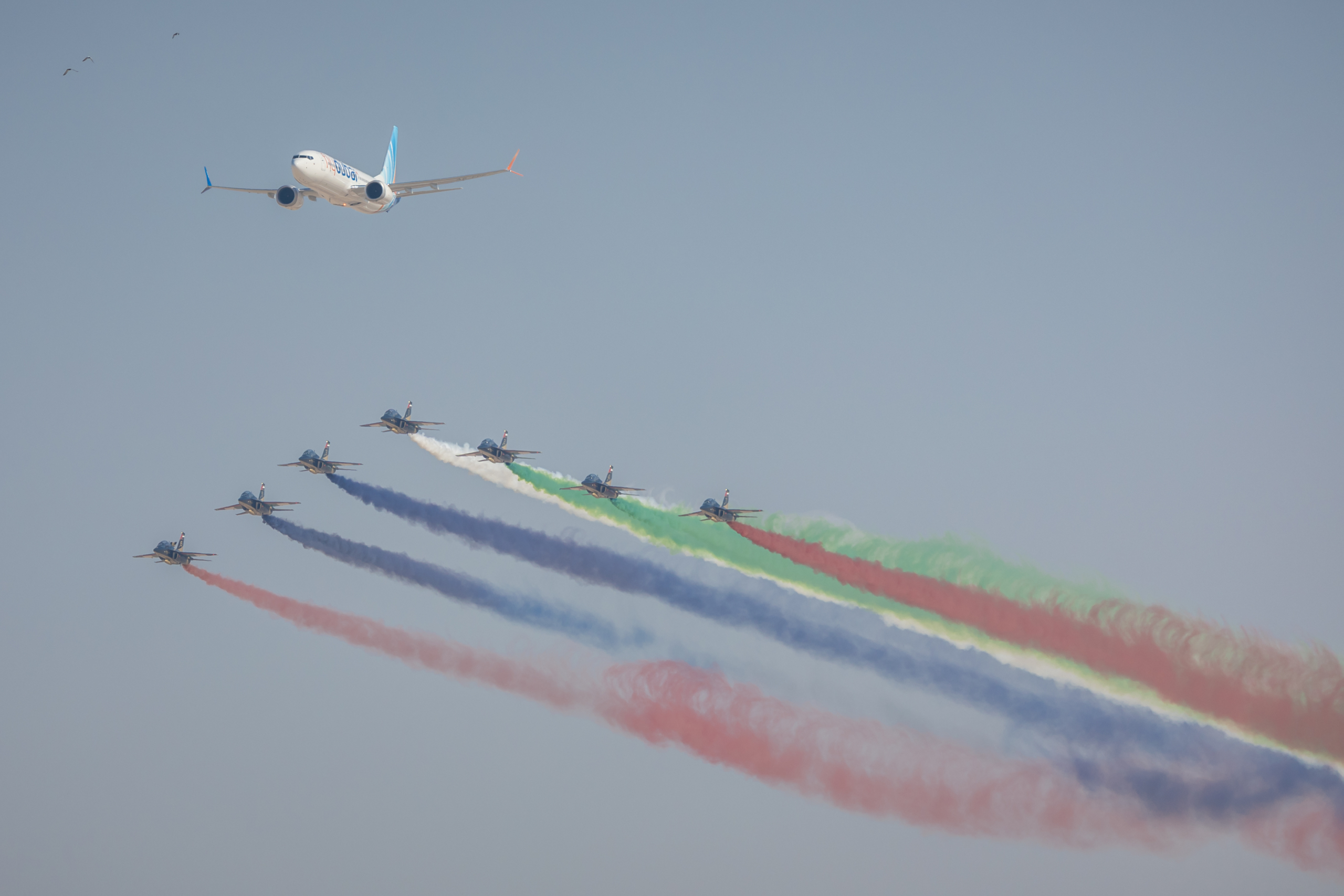Aircraft performing aerial display at Dubai Airshow with colored smoke trails and commercial airplane flying overhead