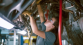 A mechanic works underneath a vehicle, using tools to perform maintenance or repairs on the car's undercarriage components.