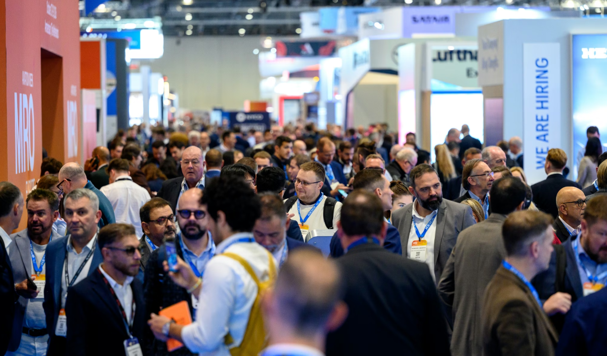 A crowded trade show or conference hall filled with attendees wearing business attire and blue lanyards, with various company exhibition booths and signage visible in the background.