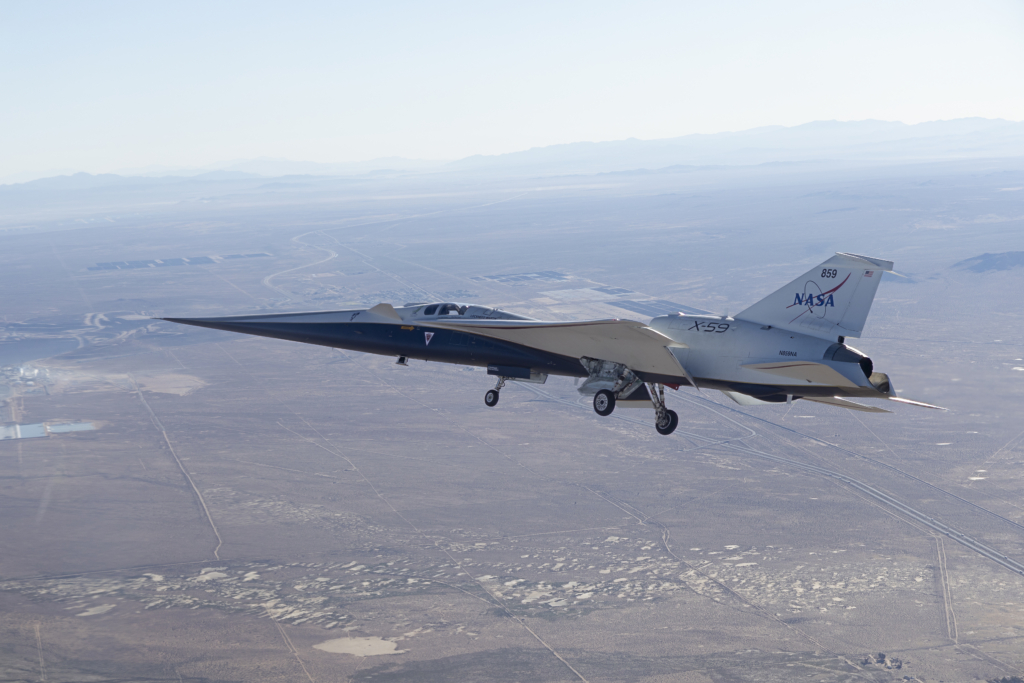 NASA’s X-59 is seen in flight, with a blue sky and mountains behind it and land below it. The aircraft’s long nose and distinct silhouette are visible as it ferries to NASA’s Armstrong Flight Research Center in Edwards, California